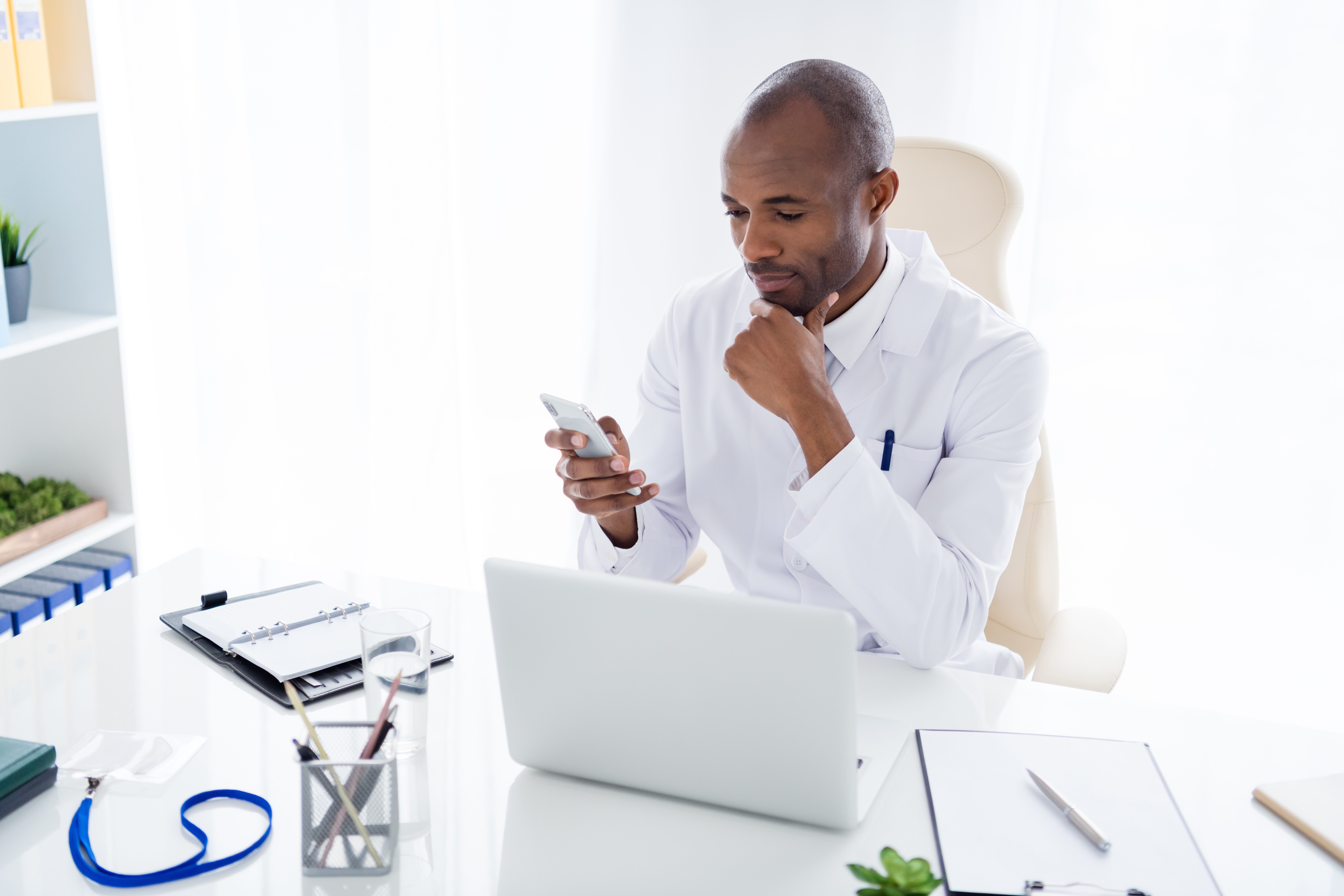 Photo of family doc dark skin guy watch notebook medical news online consultation use telephone read patients email write prescription wear white lab coat sit chair office clinic indoors